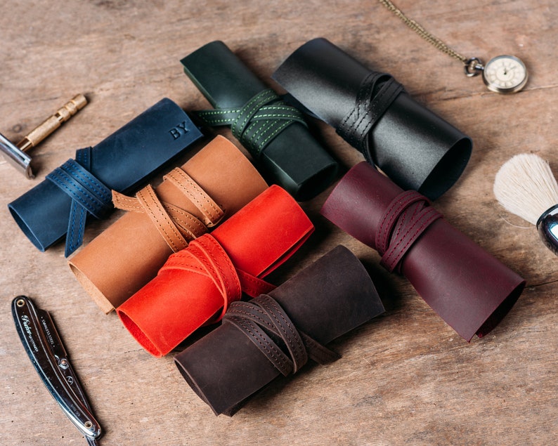 May include: Seven leather rolls in various colors, including brown, red, black, green, and blue. Each roll has a leather strap with stitching and is rolled up with a label that says "BY".
