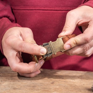 May include: A close-up of a brown leather belt with a brass buckle. The buckle is being fastened. The belt is on a wooden surface. The person's hands are visible.