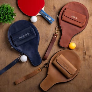 May include: A collection of leather ping pong paddle cases in navy blue, brown, and tan, each with a small wallet. One case has "SPIN TO WIN" stitched on it, and others have names. A red ping pong paddle and balls are also present.