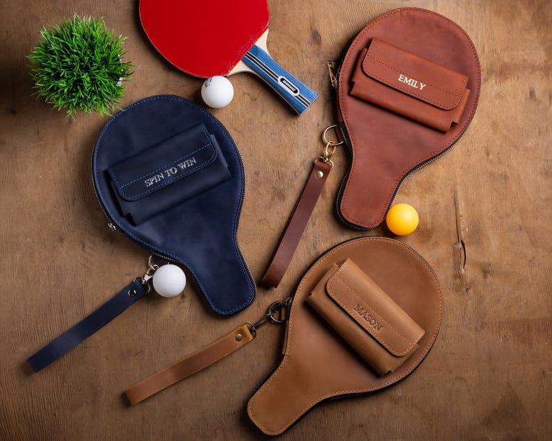 May include: A collection of leather ping pong paddle cases in navy blue, brown, and tan, each with a small wallet. One case has "SPIN TO WIN" stitched on it, and others have names. A red ping pong paddle and balls are also present.