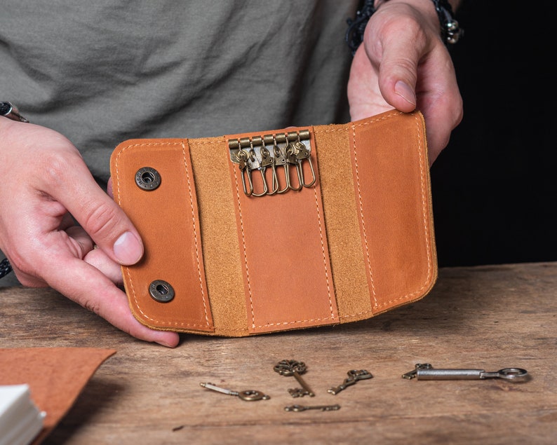 May include: A brown leather key holder with a flap closure and multiple key rings. The holder is open and the key rings are visible. The holder is on a wooden surface with several keys scattered around it.