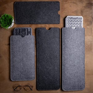 May include: A selection of grey felt keyboard sleeves and a small potted plant on a wooden surface. Two keyboards are partially visible inside the sleeves. A pair of round spectacles rests at the bottom.
