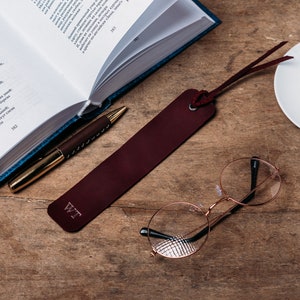 May include: A burgundy leather bookmark with the initials "WT" embossed on it. The bookmark is lying on a wooden surface next to a book, a pen, and a pair of round glasses.