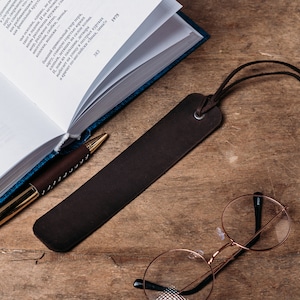 May include: A brown leather bookmark with a long tassel, a gold pen, a pair of round glasses, and a white ceramic cup on a wooden surface.