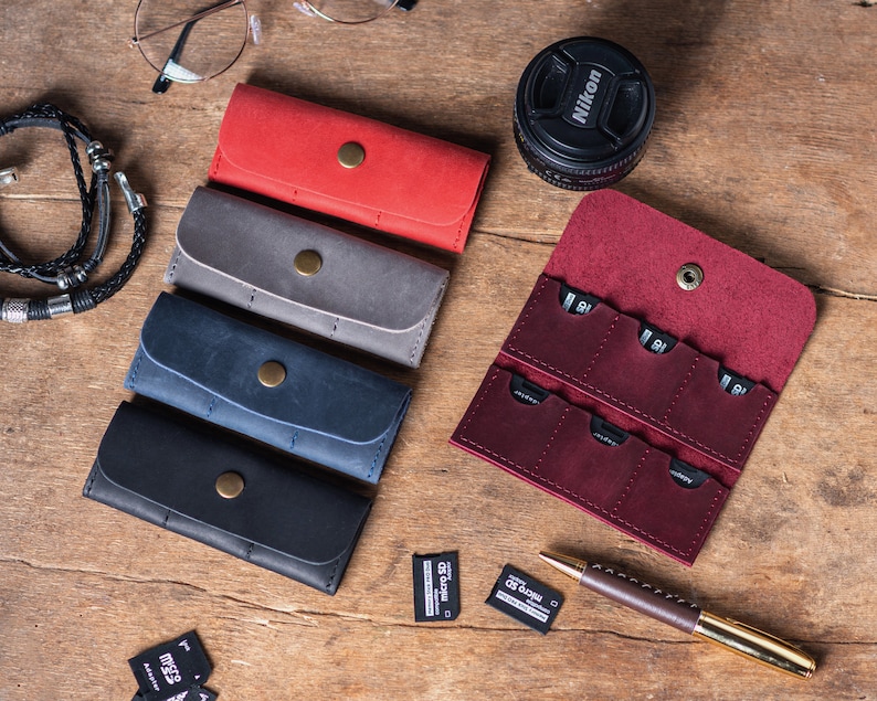 May include: An assortment of six leather cases for memory cards in various colours, including red, grey, blue, and black. The cases have a button closure and are arranged in a row on a wooden surface. A burgundy leather case with multiple slots for memory cards is open and visible in the background.