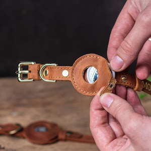 Op de afbeelding: Een bruine leren hondenriem met een ronde, afneembare leren tag die een metalen drukknopsluiting heeft. De tag heeft een klein, transparant venster. De riem is gegraveerd met het woord "Mokey".