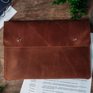 Puede incluir: Una carpeta de cuero marrón con dos broches metálicos. La carpeta está sobre una superficie de madera con papeles y útiles de escritura cerca.