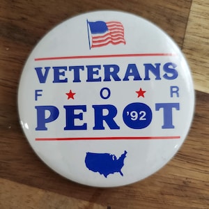 May include: A white button with blue and red text that reads "Veterans For Perot '92". The button also features a small American flag in the top right corner and a blue outline of the United States in the bottom center.