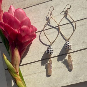 May include: Gold-toned, diamond-shaped earrings with dangling seashell and pearl accents. The earrings are displayed on a white wooden surface next to a vibrant pink flower. The earrings have a delicate, handcrafted appearance.
