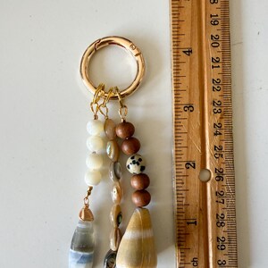 May include: A gold-toned metal keyring featuring three dangling strands of beads. The strands include white, brown, and spotted beads, along with a gray-blue teardrop stone and a large tan cone shell. A wooden ruler shows the item is 3 inches long.