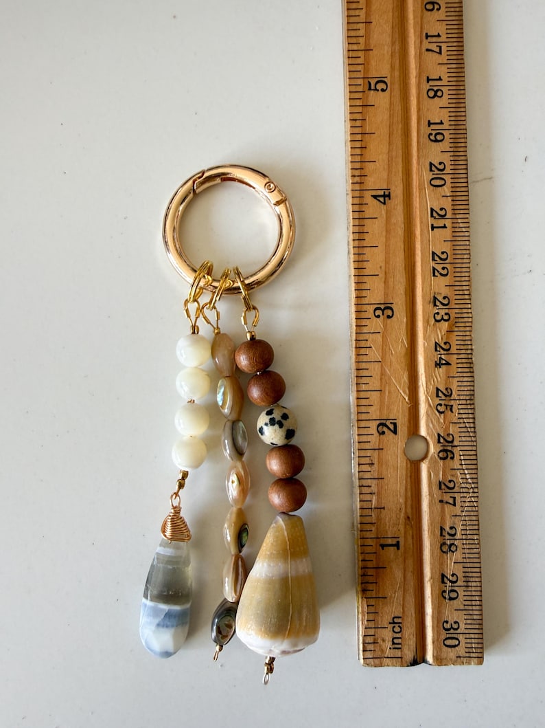 May include: A gold-toned metal keyring featuring three dangling strands of beads. The strands include white, brown, and spotted beads, along with a gray-blue teardrop stone and a large tan cone shell. A wooden ruler shows the item is 3 inches long.