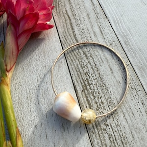 May include: A gold-toned bangle bracelet featuring a natural seashell and a small, round, gold-colored bead. The bracelet rests on a weathered, light gray wooden surface, with a vibrant pink flower in the background.