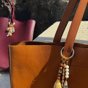 May include: A tan leather tote bag featuring a gold-toned metal ring attachment adorned with a multi-strand charm. The charm includes brown wooden beads, a spotted bead, white spherical beads, and two distinct dangling shell-like pendants.