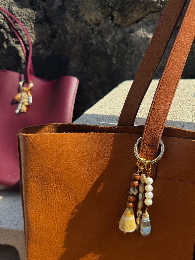 May include: A tan leather tote bag featuring a gold-toned metal ring attachment adorned with a multi-strand charm. The charm includes brown wooden beads, a spotted bead, white spherical beads, and two distinct dangling shell-like pendants.