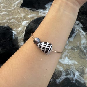 May include: A gold-toned metal bangle bracelet featuring a single dark grey spherical pearl and a white seashell with a distinct black spotted pattern. The bracelet is a simple open-ended design that rests on the forearm, showcasing the contrast between the metallic band, the smooth pearl, and the textured natural shell.