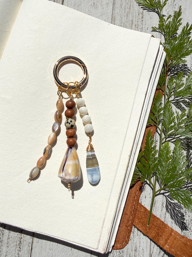 May include: A gold-toned metal keyring featuring three dangling charms. One strand has oval-shaped iridescent beads, another has brown wooden beads with a spotted stone, and a large conical shell, while the third has white beads and a teardrop-shaped stone.