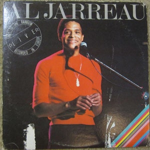 Op de afbeelding: Een zwart-wit albumhoes met Al Jarreau, een zanger, die een rode trui en een zilveren ketting draagt. De albumtitel is "Look to the Rainbow - Live - Recorded in Europe".
