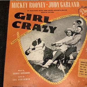 May include: A vintage record album cover featuring Mickey Rooney and Judy Garland in a scene from the musical "Girl Crazy". The cover is orange with a yellow rope border and features the title "Girl Crazy" in large black letters. The album is by Decca Records.