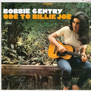 May include: A woman with long brown hair sits on a wooden fence, playing a brown acoustic guitar. She is wearing a white t-shirt and blue jeans. The album cover reads "Bobbie Gentry Ode to Billie Joe" with a Capitol Records logo.