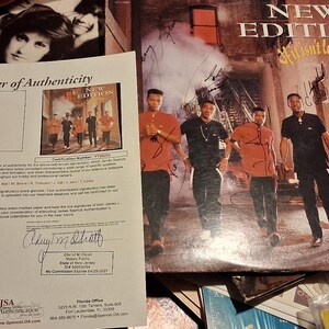 May include: A signed album cover for New Edition's "If It Isn't Love" album. The cover features the band members in red and black shirts. A letter of authenticity is in the foreground, with the JSA Authentication logo.