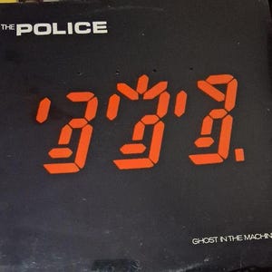 May include: Black album cover with the band name "THE POLICE" in white at the top. Large orange digital-style numbers and symbols are in the center. The album title "GHOST IN THE MACHINE" is at the bottom.