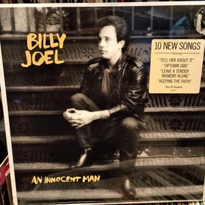 Può includere: Una foto in bianco e nero di Billy Joel seduto su una scalinata. La copertina dell'album è per il suo album "An Innocent Man" e presenta il titolo dell'album in testo giallo. La copertina dell'album include anche una scatola gialla con il testo "10 New Songs" e un elenco delle canzoni incluse nell'album.