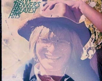 May include: A vintage album cover for John Denver's Greatest Hits. The cover shows a smiling person wearing a hat and glasses. The album title is in green text.