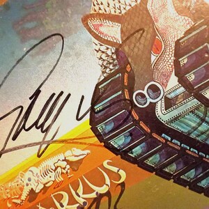 May include: Close-up of a vintage album cover with a signature in black ink. The artwork features a stylized creature with a reptilian eye and a tank-like structure. The color palette includes shades of blue, orange, and brown.