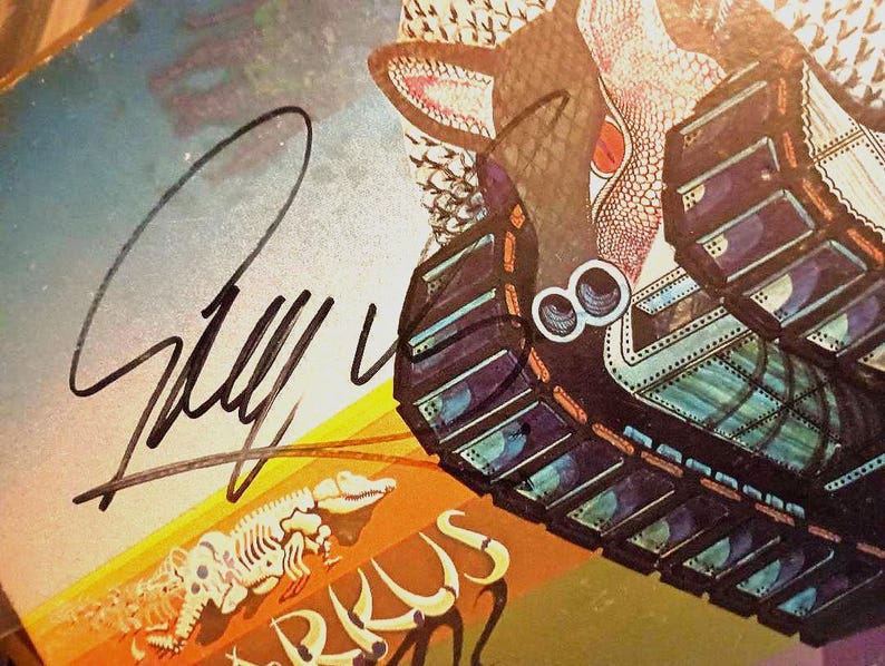 May include: Close-up of a vintage album cover with a signature in black ink. The artwork features a stylized creature with a reptilian eye and a tank-like structure. The color palette includes shades of blue, orange, and brown.