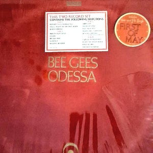 Puede incluir: Un álbum de vinilo rojo de los Bee Gees titulado "Odessa". La portada presenta letras doradas y una pegatina que dice "First of May". Una lista de canciones está impresa en una etiqueta blanca. Es un álbum doble.