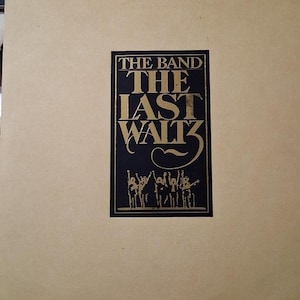 May include: A tan album cover with a black and gold rectangular graphic. The graphic reads "THE BAND THE LAST WALTZ" with an illustration of musicians. The album cover is slightly worn.