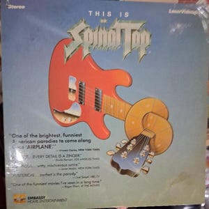 May include: Album cover for "This Is Spinal Tap" with a red guitar graphic. The design includes quotes from critics and the Embassy Home Entertainment logo. The background is a blue gradient. The text reads "This Is Spinal Tap".
