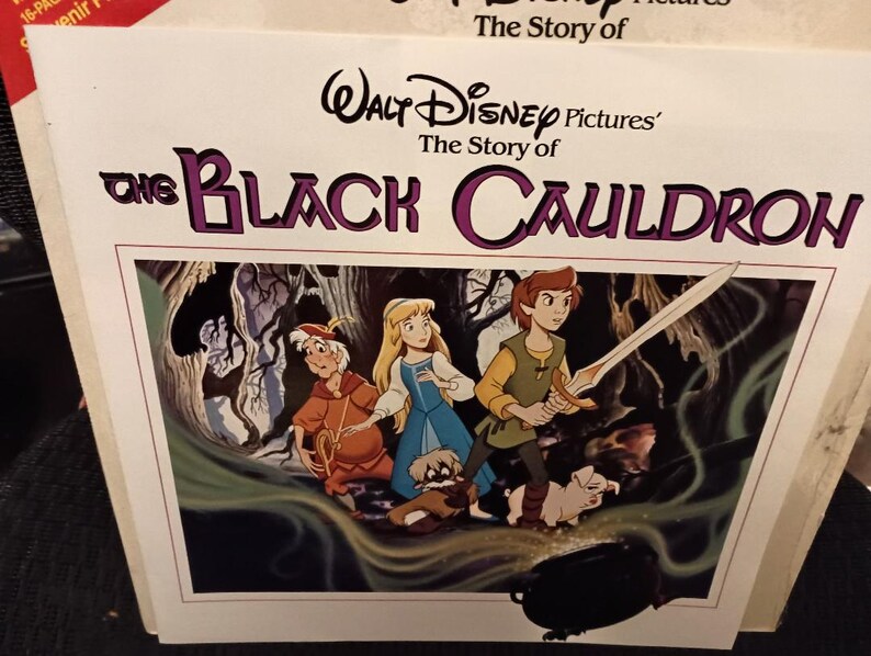 May include: A book cover for "The Black Cauldron" featuring a colorful illustration of characters from the Disney film. The title is in large, purple letters. The image shows a young man with a sword, a woman in a blue dress, and other characters.