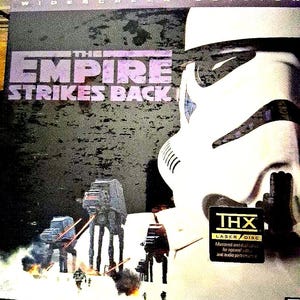 May include: A vintage LaserDisc of "The Empire Strikes Back" featuring a Stormtrooper helmet and AT-AT walkers. The cover has the film's title in purple and the THX LaserDisc logo.