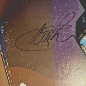 May include: Close-up of a vintage album cover with a signature in black ink. The cover has a gradient of brown, purple, and blue hues. The signature is stylized and appears to be a musician's autograph.