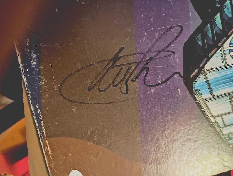 May include: Close-up of a vintage album cover with a signature in black ink. The cover has a gradient of brown, purple, and blue hues. The signature is stylized and appears to be a musician's autograph.