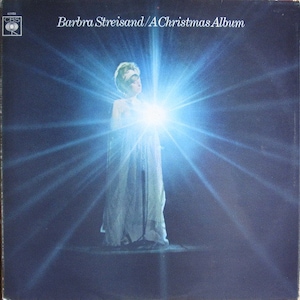 Puede incluir: Una mujer con un vestido blanco se encuentra frente a una luz azul brillante, con un efecto de estrella que irradia desde la luz. El texto "Barbra Streisand/A Christmas Album" está impreso en la parte superior de la imagen.