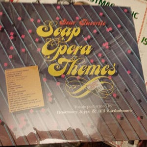 May include: A vinyl record album titled "Your Favorite Soap Opera Themes." The album cover is dark blue with diagonal lines and pink and red accents. The title is in large yellow script. The album features vocals by Rosemary Joyce & Bill Bartholomew.