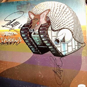 May include: Album cover art featuring a surreal illustration of an armadillo-like creature in a tank. The artwork is signed and includes the text "Tarkus" in a stylized font. The color palette includes blues, browns, and purples.
