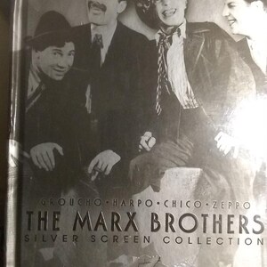 Puede incluir: Una caja de DVD plateada con una foto en blanco y negro de los hermanos Marx. El texto en la caja dice "Groucho, Harpo, Chico, Zeppo The Marx Brothers Silver Screen Collection".