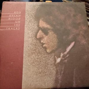May include: Album cover for Bob Dylan's "Blood on the Tracks." The album art features a profile of a person with sunglasses against a textured background. The album title and artist's name are printed in white on a maroon vertical stripe.