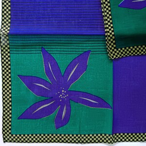May include: A colorful textile featuring a large purple flower with white accents on a teal green background. The design includes a blue section with black horizontal lines and a checkerboard border in black and tan.