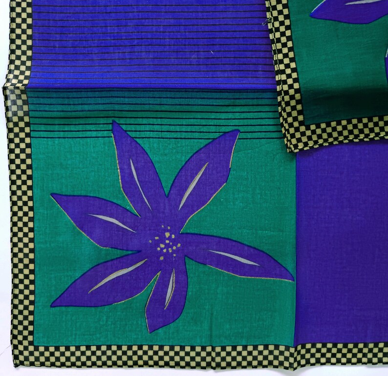 May include: A colorful textile featuring a large purple flower with white accents on a teal green background. The design includes a blue section with black horizontal lines and a checkerboard border in black and tan.
