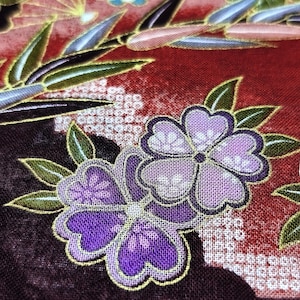 May include: A fabric featuring a deep red background with a pattern of purple flowers and green bamboo leaves. The flowers have five petals outlined in gold, and the bamboo leaves are accented with gold edges. White geometric patterns are visible.