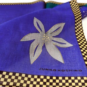 May include: A folded blue scarf with a large, stylized gray flower with gold accents. The scarf has a checkered border in black and yellow, and the text "JUNKO KOSHINO" is printed on the fabric.