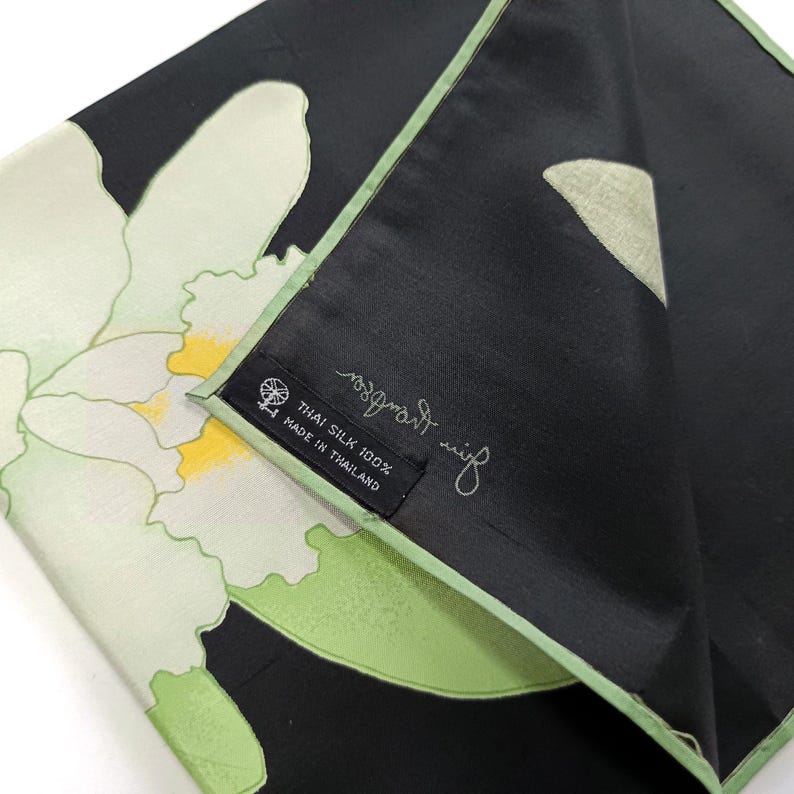 Puede incluir: Pa&ntilde;uelo de seda negro con un dise&ntilde;o floral con flores blancas y amarillas con detalles en verde. El pa&ntilde;uelo tiene un borde verde y una etiqueta que dice "Thai Silk 100% Made in Thailand."