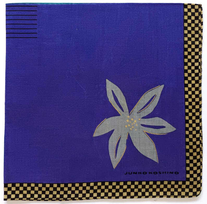 May include: Square blue handkerchief with a gray floral design and a black and tan checkered border. The handkerchief has the text "JUNKO KOSHINO" printed on it.