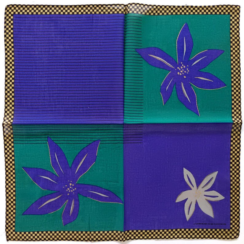 May include: A square, folded textile with a checkered border. The design features four quadrants: two blue with horizontal lines, and two teal with purple floral designs. A white floral design is in the lower right quadrant.