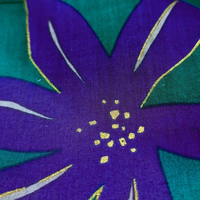 May include: Close-up of a fabric with a large, stylized purple flower design. The flower has white and gold accents, set against a green background. The design is likely part of a textile or decorative item.