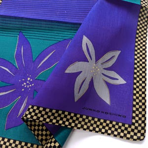 May include: A folded square textile with a floral design. The textile features a large purple flower with silver accents on a teal background. A second section is purple with a silver flower. The border is black and tan checkered. The text "JUNKO KOSHINO" is visible.